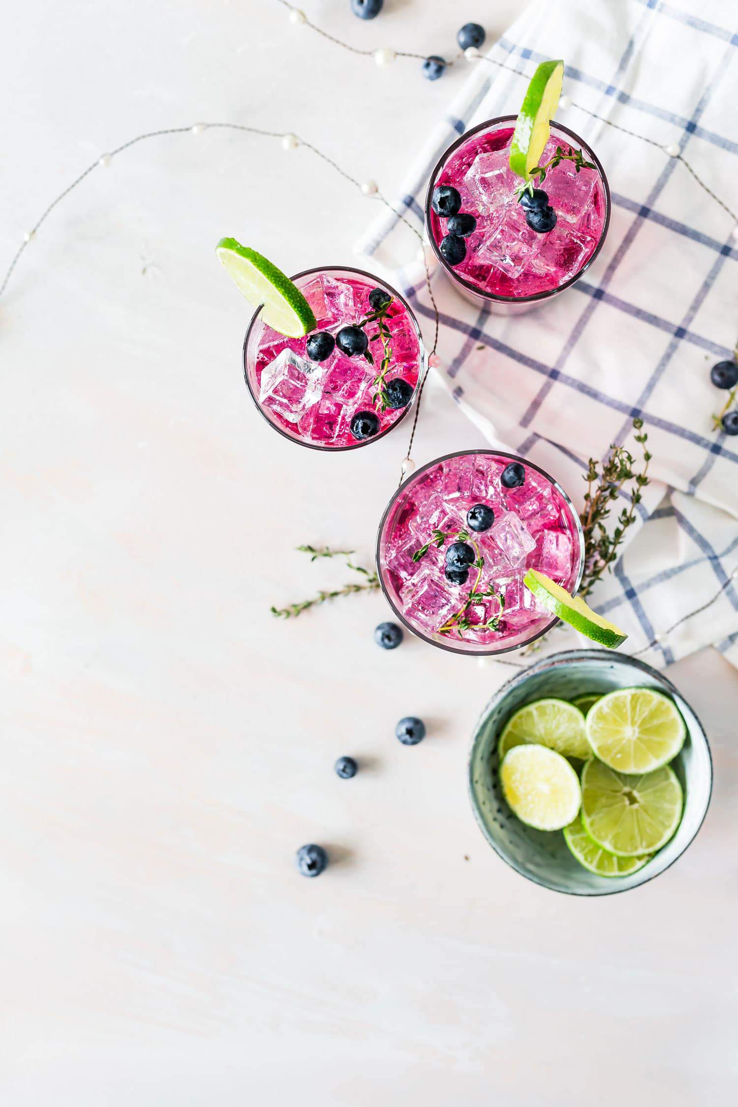 three glasses of blueberry lime refresher bowl of limes blue and white checkered towel drinks garnished with thyme