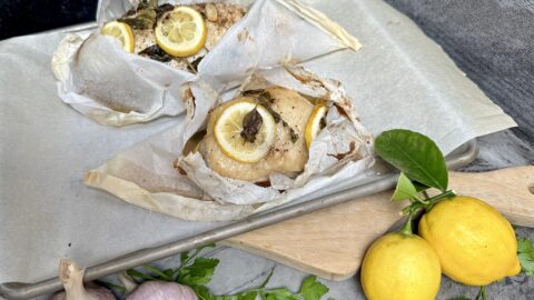 Chicken En Papillote chicken in parchment paper package on a sheet pan garnished with lemon garlic herbs