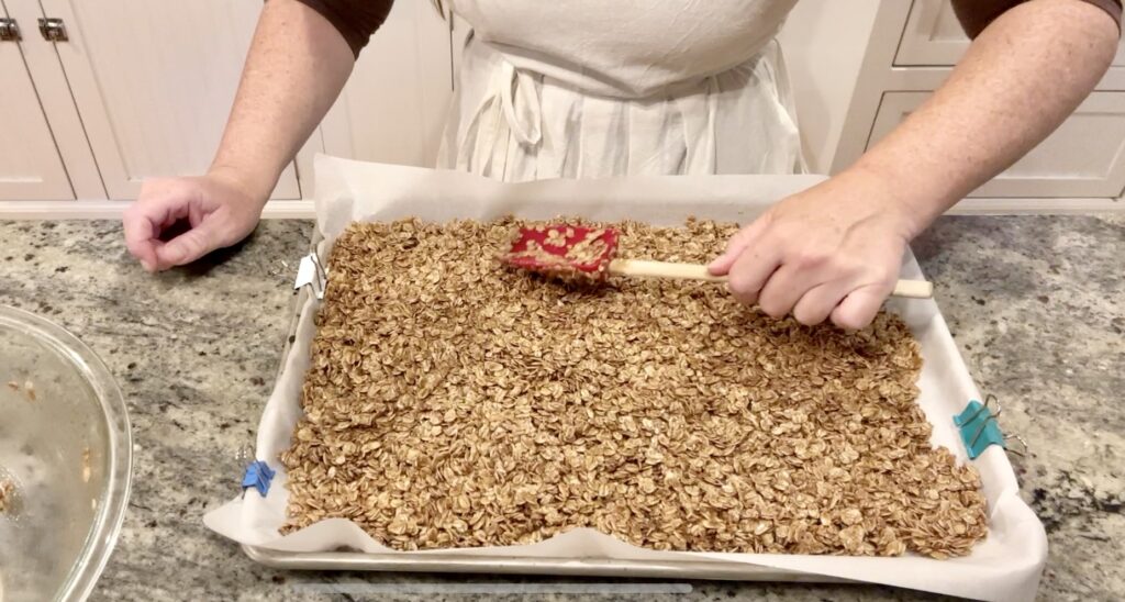 woman in a white apron standing behind cinnamon pepita granola and spreading it on sheet pan covered in parchment