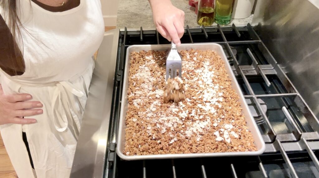 women in white apron stiring coconut in granola on a half sheet pan with a stainless steel spatula on a cook top
