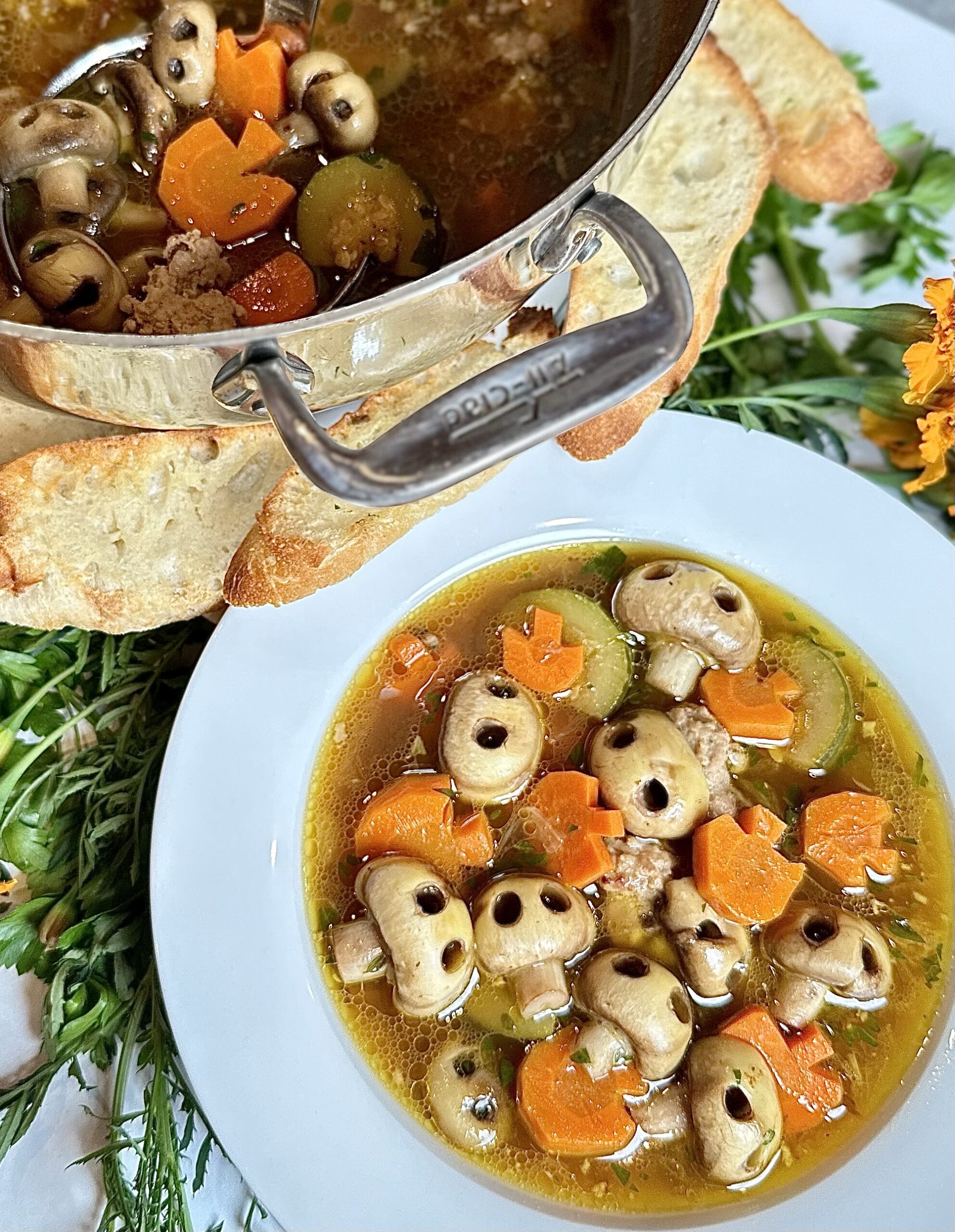 Spooky Sausage Soup with mushroom skulls in an all clad Dutch oven served in a white rimmed soup bowl arnished with herbs carrots that look like pumpkins