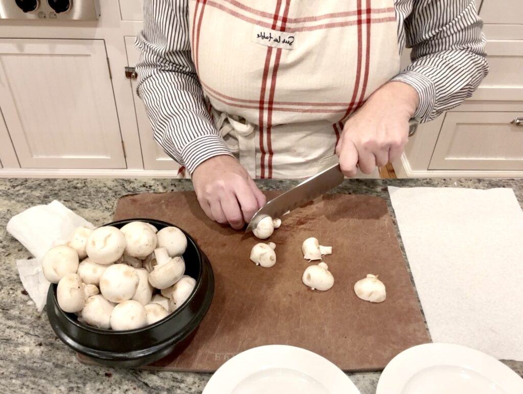 woman in an apron is cutting a mushroom in half on a cutting board