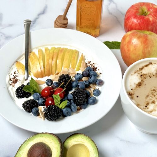 labne breakfast bowl with apple blackberries blueberries cherries sliced almonds latte in a white mug honey cocoa powder sliced avocado to garnish