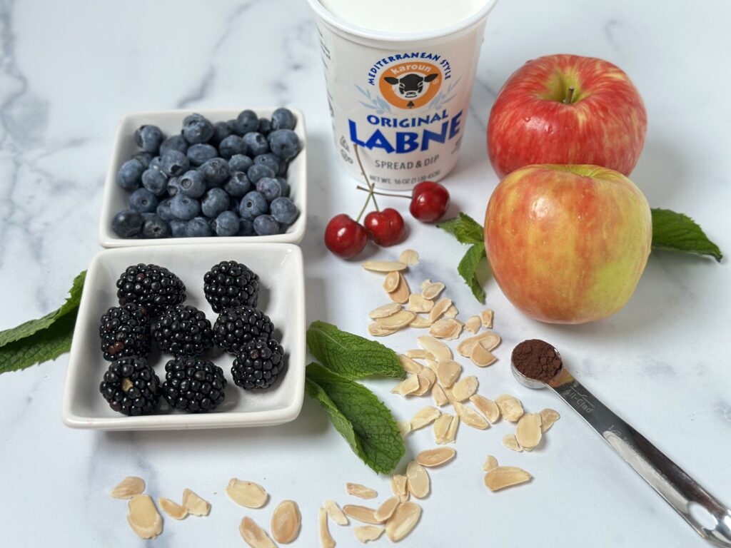 labne breakfast bowl with fruit ingredients apple blueberries blackberries cocoa powder in a measuring spoon sliced almonds cherries mint leaf labne in a container on a white marble counter top