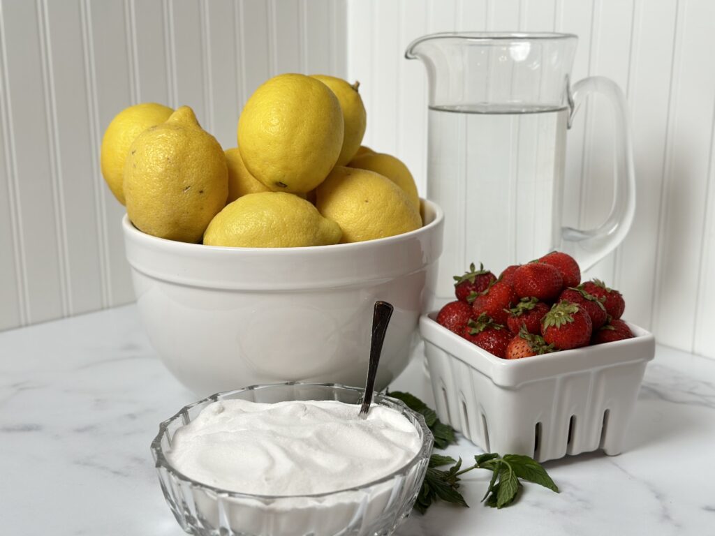 strawberry lemonade ingredients white bowl with lemons glass bowl with sugar white ceramic fruit basket with strawberries glass pitcher with water