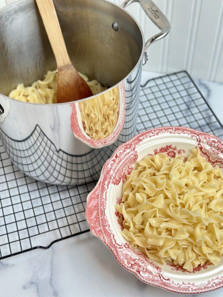 buttered noodles in an All-Clad Pasta pot on a cooling rack next to a bowl of buttered noodles in a Johnson Brothers Old Britain Castles Pink & Cream red and white covered vegetable dish