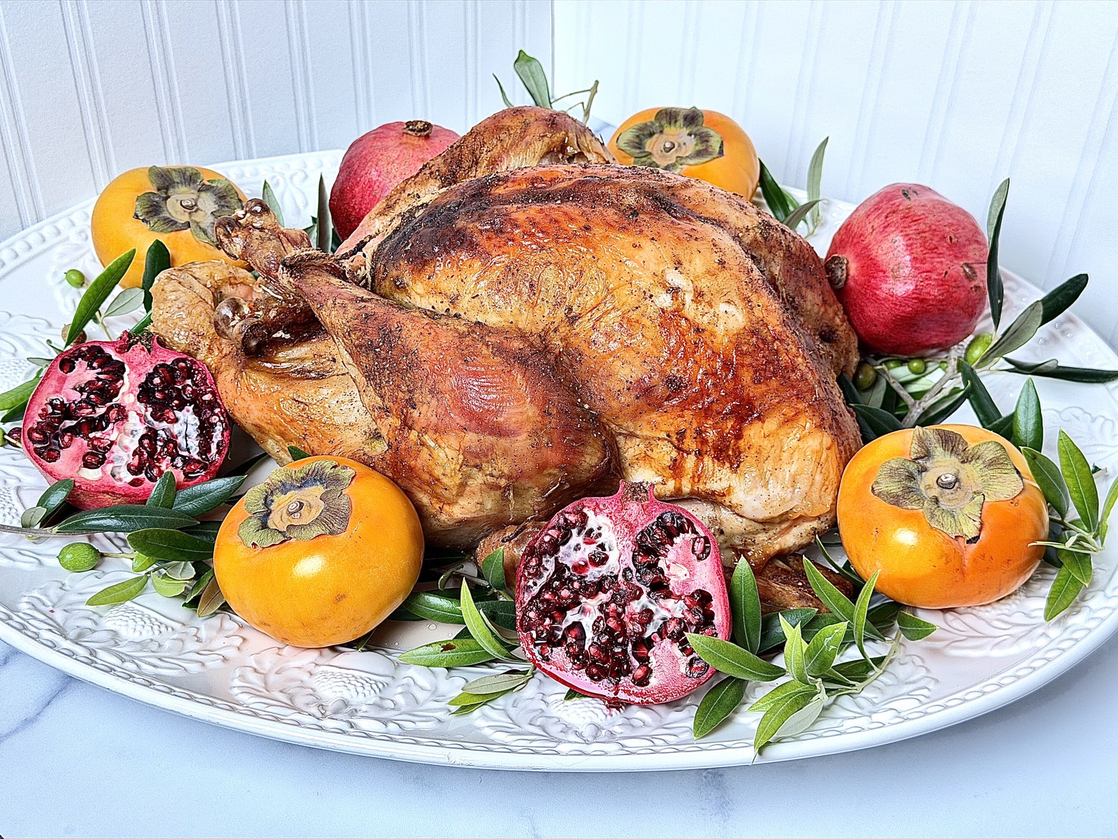 Image of the perfect turkey on a white serving platter with acorns, olive branches, pomegranates, and persimmons on a marble counter