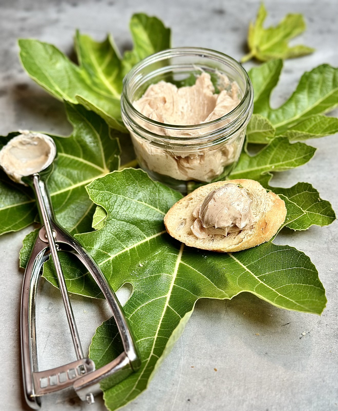 Picture of an ice cream scooper putting a dollop of Cinnamon, honey, compound butter from A jar on top of a sliced baguette, on top of fig leaves
