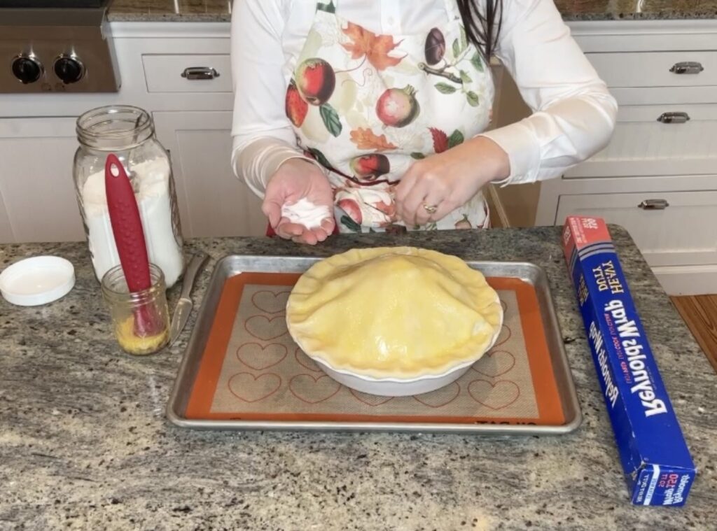 A woman in a white shirt and fall apron on making a homemade apple pie and is sprinkling on the top crust after the egg wash was added.