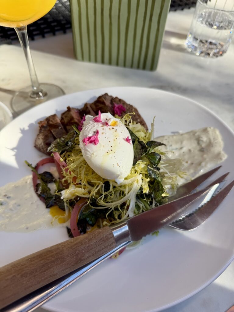 a picture of steak and eggs from the Clementine Yountville restaurant, it is sliced filet, baby, heirloom, tomatoes, watercress, pickled red onion salad with horseradish cream, and a poached egg on a white plate with a knife and fork on the white plate also