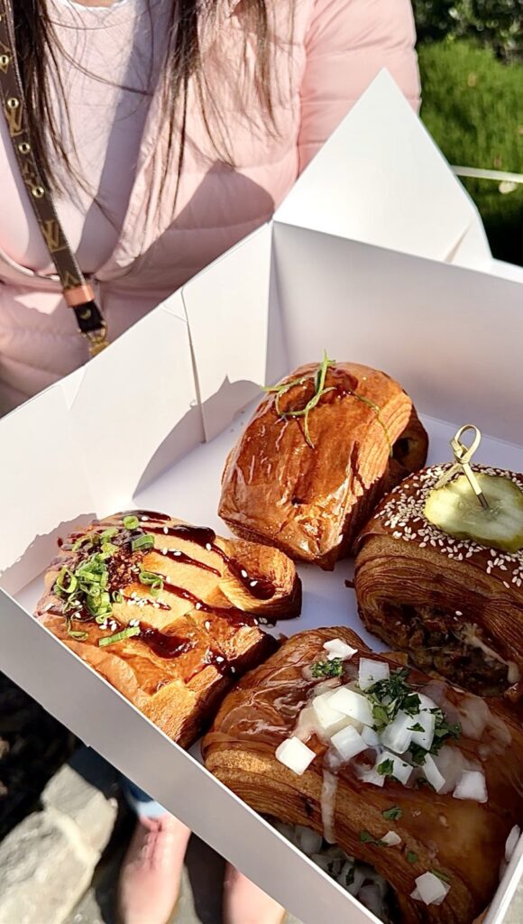 A picture of a woman in pink Doc, Martens and a pink Lululemon jacket holding a box of savory pastry from butter and crumble in San Francisco, standing in Washington Park.