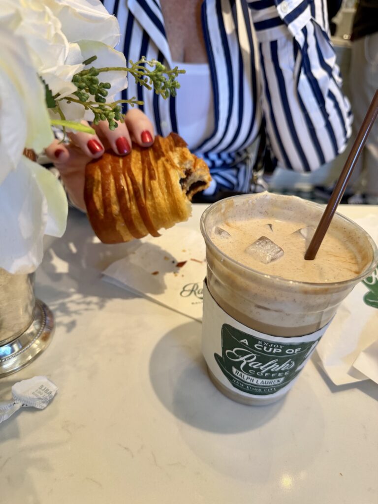 A woman in a blue and white striped Ralph Lauren dress, holding a pawn of shock a lot with one bite out of it and a iced latte with cinnamon brown sugar cold foam on a marble table