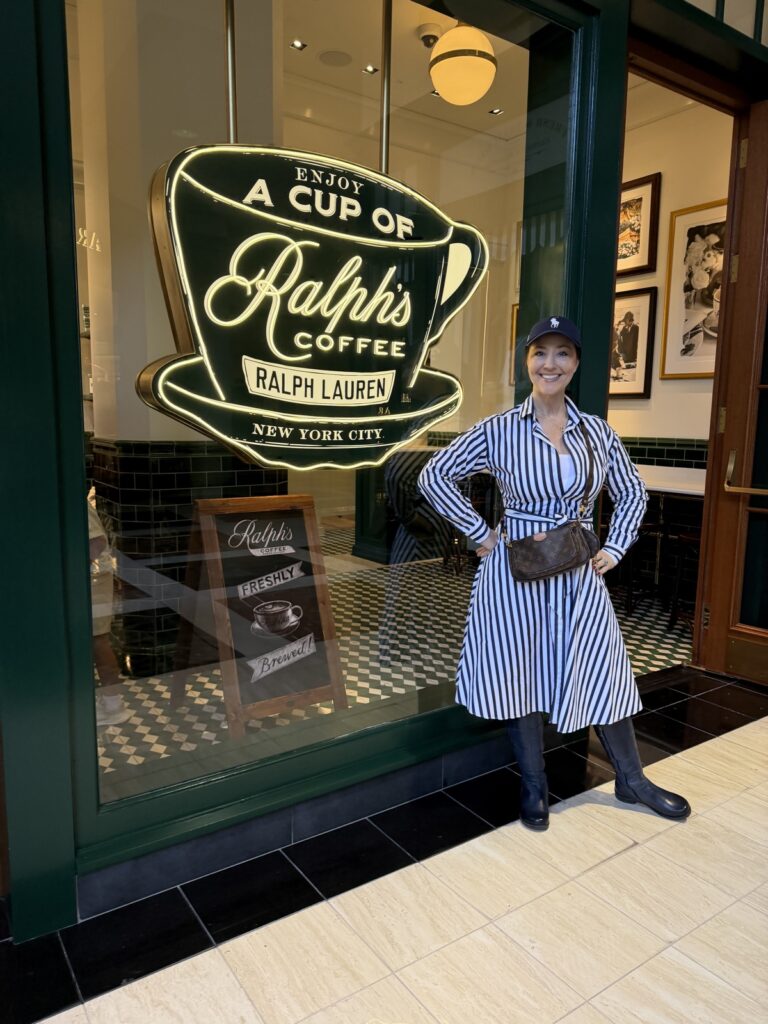 A woman in a blue striped polo, Ralph Lauren dress wearing blue leather boots and a blue polo hat with a white polo horse is standing in front of Ralph's coffee next to the iconic Ralph coffee Ralph Lauren sign