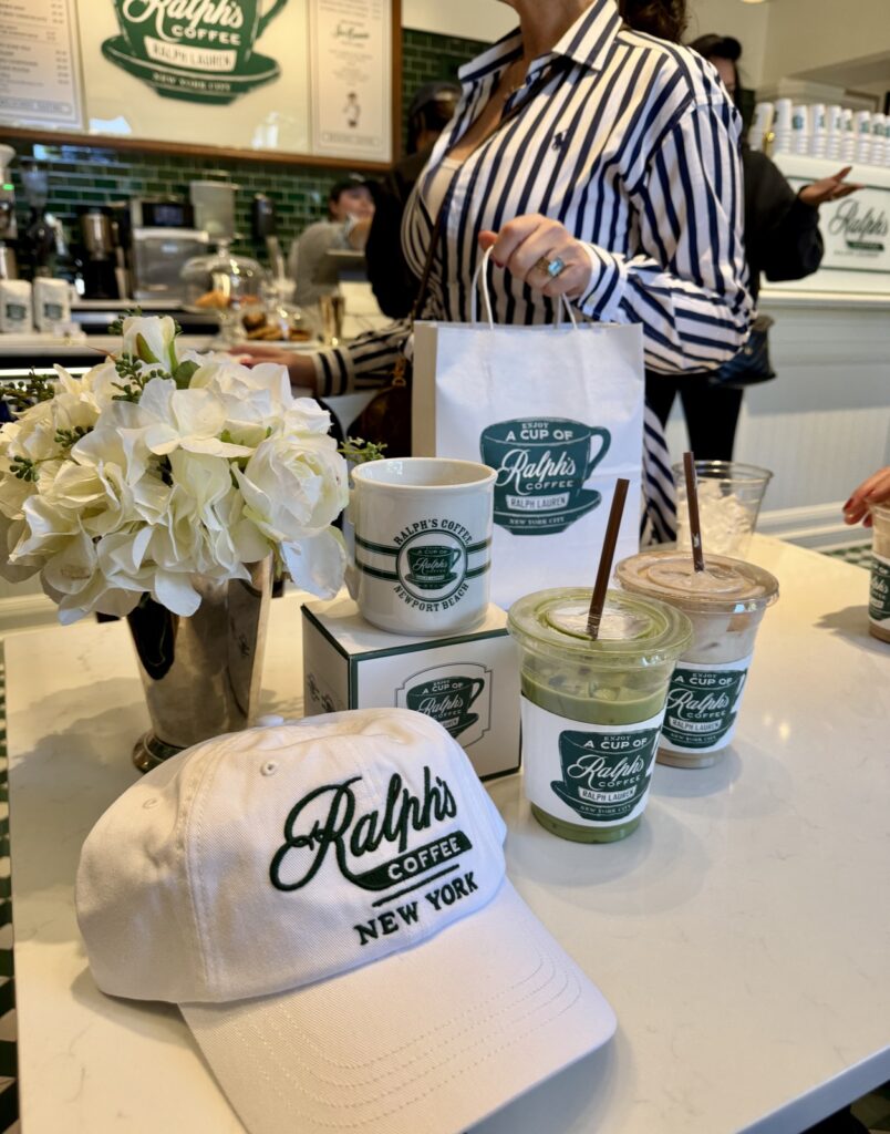 A picture of a woman wearing a blue and white striped Ralph Lauren dress holding a Ralph's coffee bag behind a Ralph's coffee, New York hat, two lattes and a Ralph's coffee Newport Beach mug