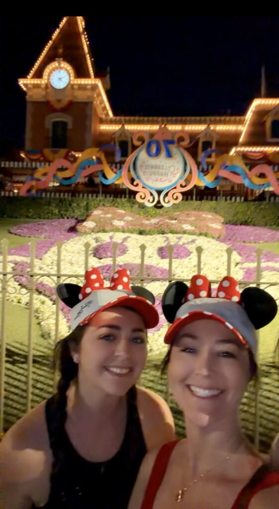 A picture of two women in Minnie mouse baseball caps in front of the Disneyland train station, decorated for Disneyland 70th anniversary celebration