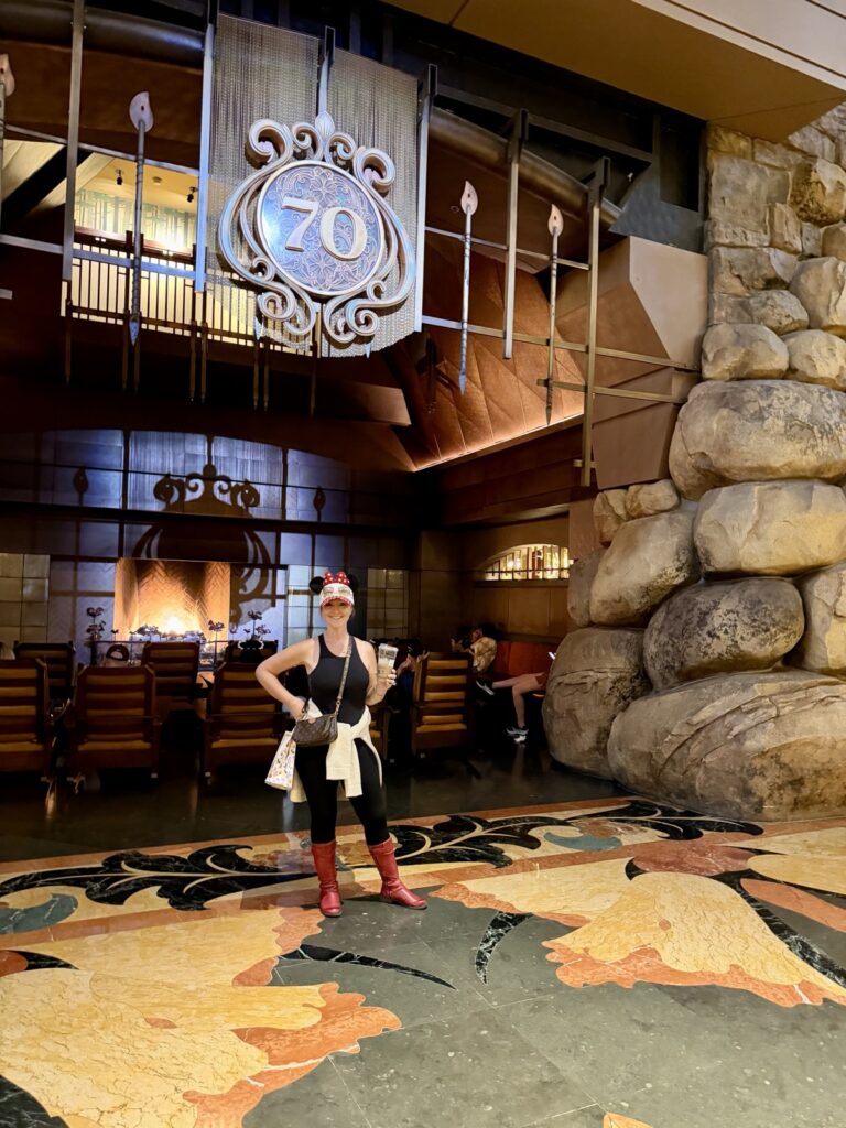 This is a picture of Rachael from a life full of living standing in the grand Californian hotel at Disneyland resort standing underneath a 70th decoration beneath the hearth of the fireplace area in the lobby
