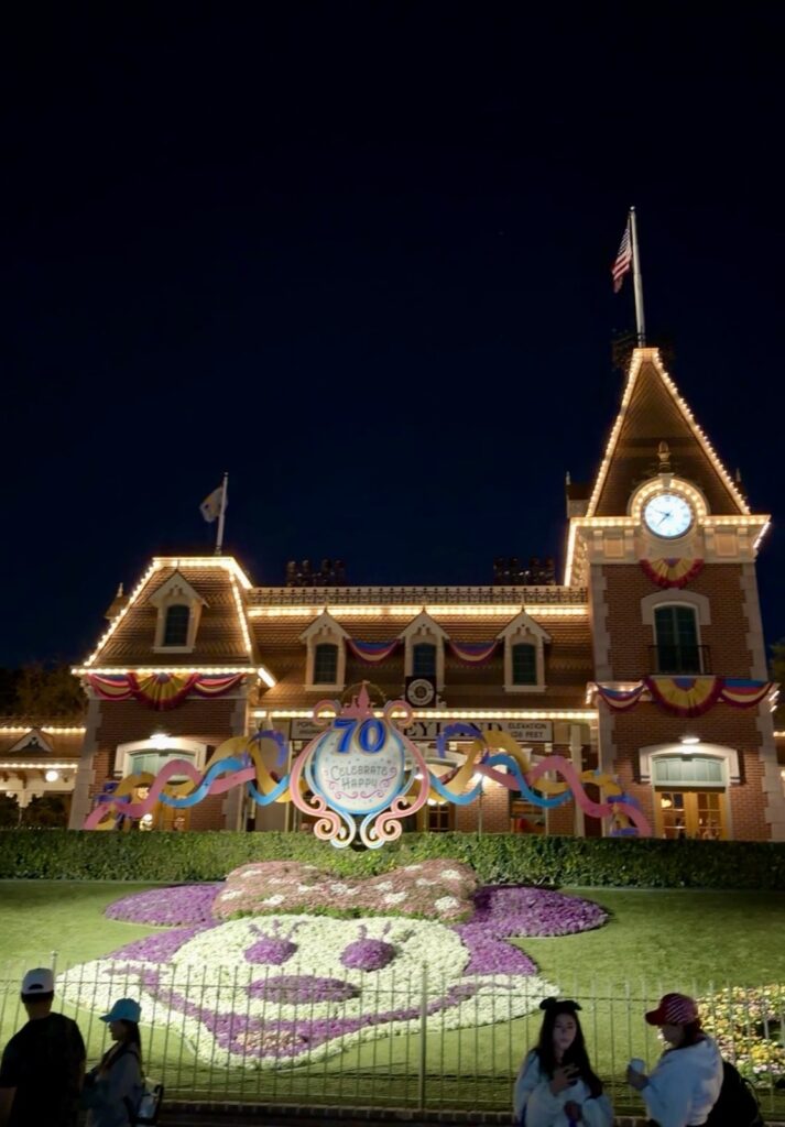 This is a picture of Disneyland train station entrance right after you enter the gates. It shows a 70th birthday celebration decoration and a beautiful Minnie mouse created with flowers with a well-maintained grass backdrop.