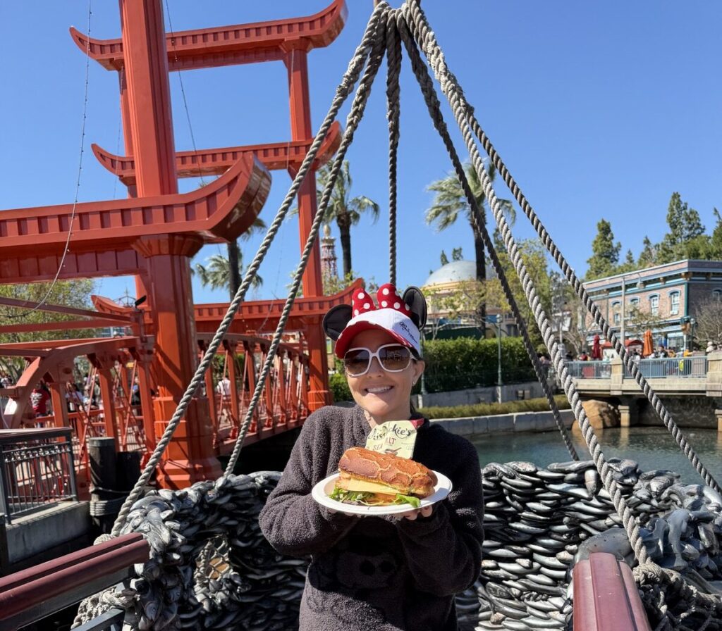 A picture of a woman in a Mickey sweatshirt and a Minnie mouse baseball cap wearing white sunglasses holding a turkey sandwich on Dutch crunch from Aunt Cass Café in Disneyland California Adventure.