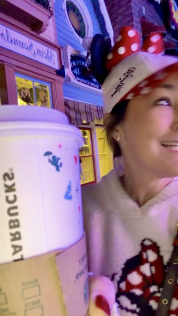 A picture of a woman wearing a Mickey hat and a Mickey sweatshirt holding a Disney Starbucks cup on Main Street USA at Disneyland.