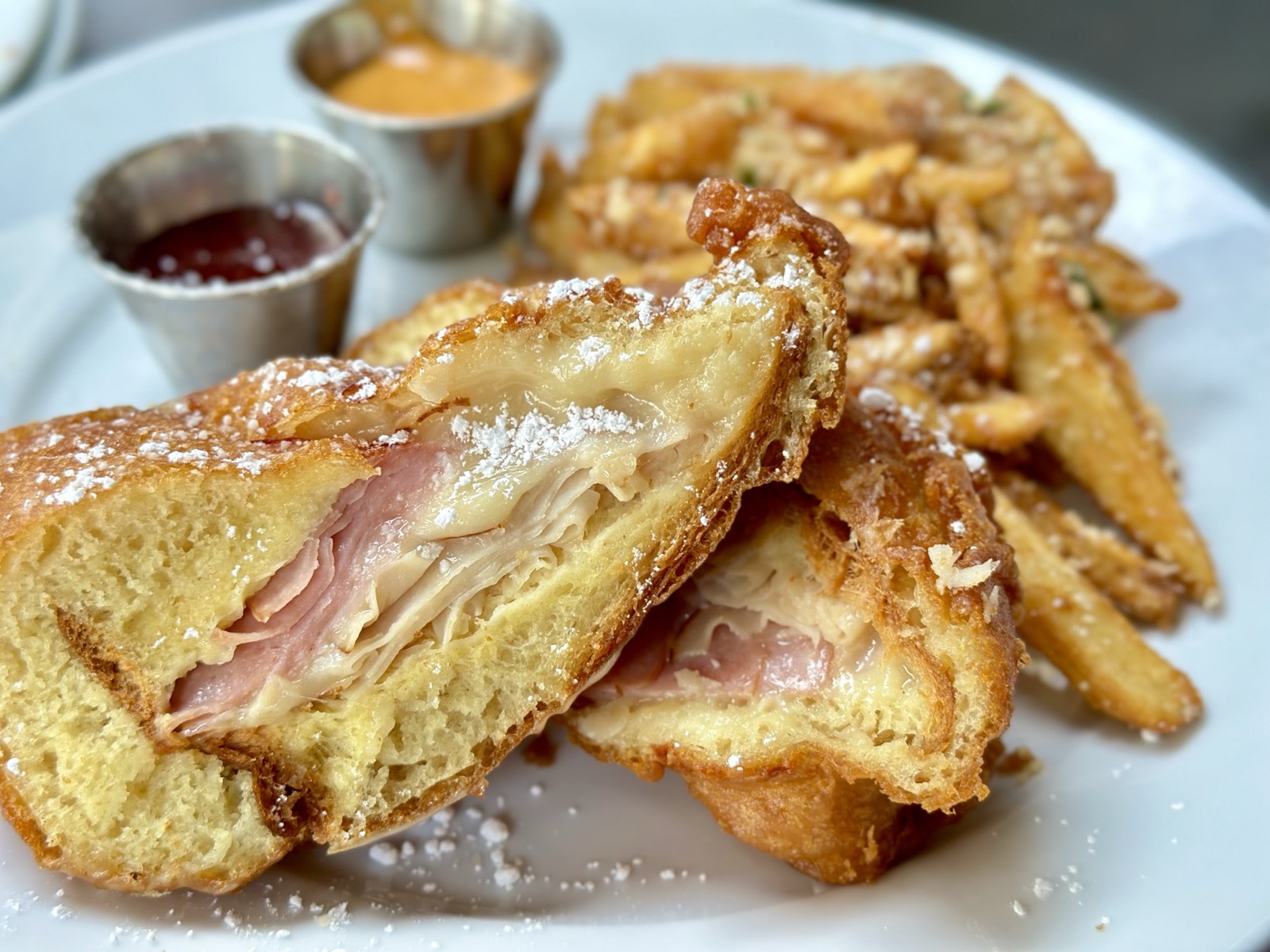 A picture of Disneyland Montecristo from Café Orleans, a sandwich made with ham, turkey, Swiss cheese, then battered and deep-fried, dusted with powdered sugar and served with served with a side of seasonal jam, and pommes frites with a creole aïoli for dipping the pommes frites in.