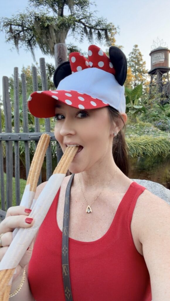 A picture of a woman wearing a Minnie mouse, baseball cat and a red tank top eating a churro.