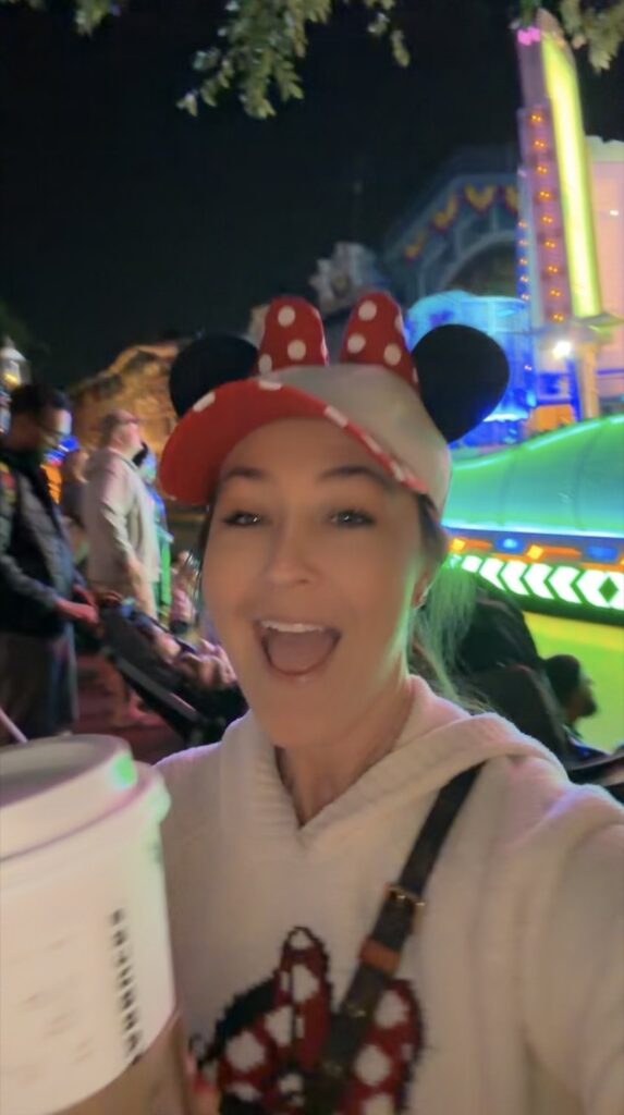 A picture of a woman wearing a Mickey Mouse hat, holding a Disney Starbucks cup with the Disney parade behind her.