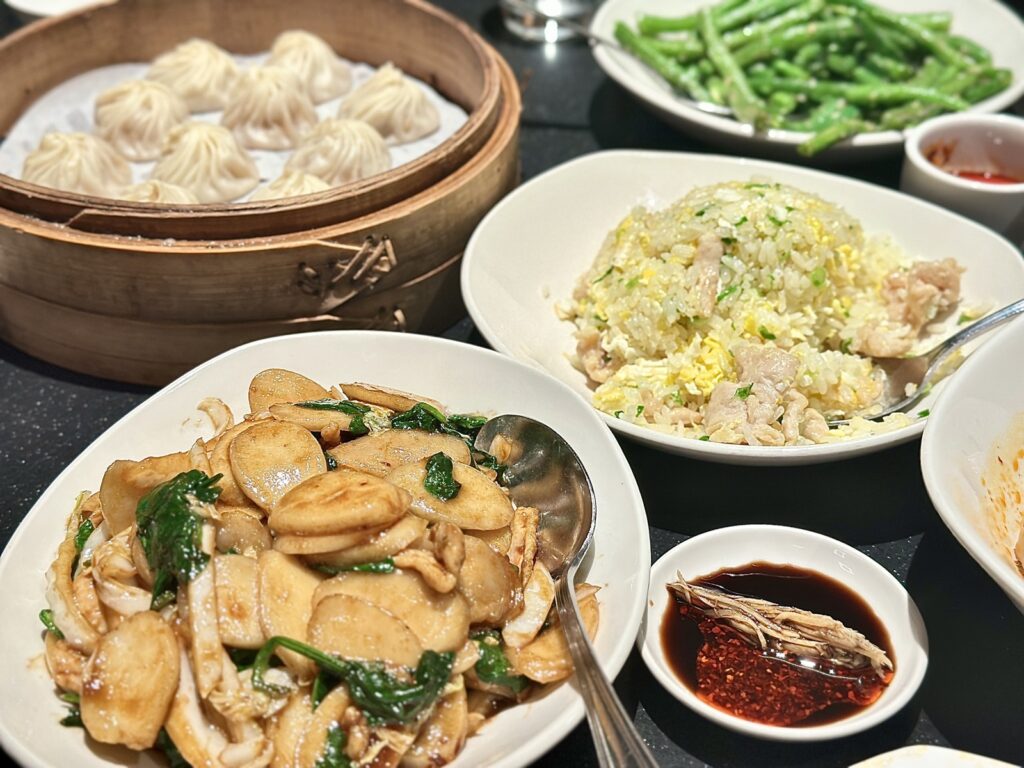 A picture of a table full of food from Din Tai Fung featuring rice, cakes, chicken, fried rice, green beans, soup dumplings, and sauces.