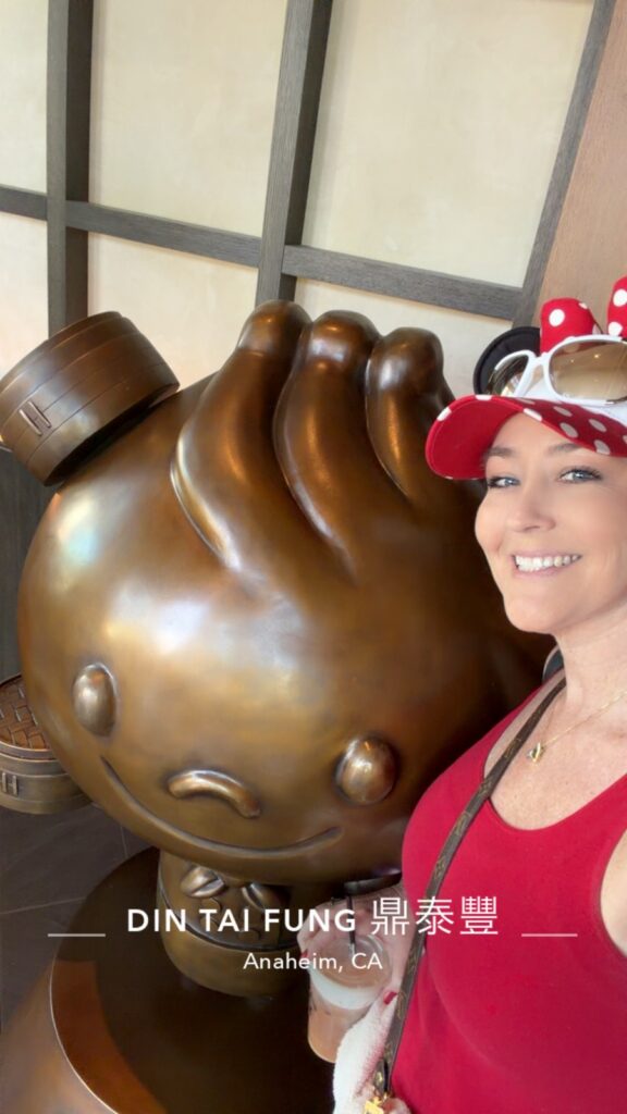 A picture of a women wearing a Minnie Mouse hat at Din Tai Fung Anaheim standing next to the dumpling statue.