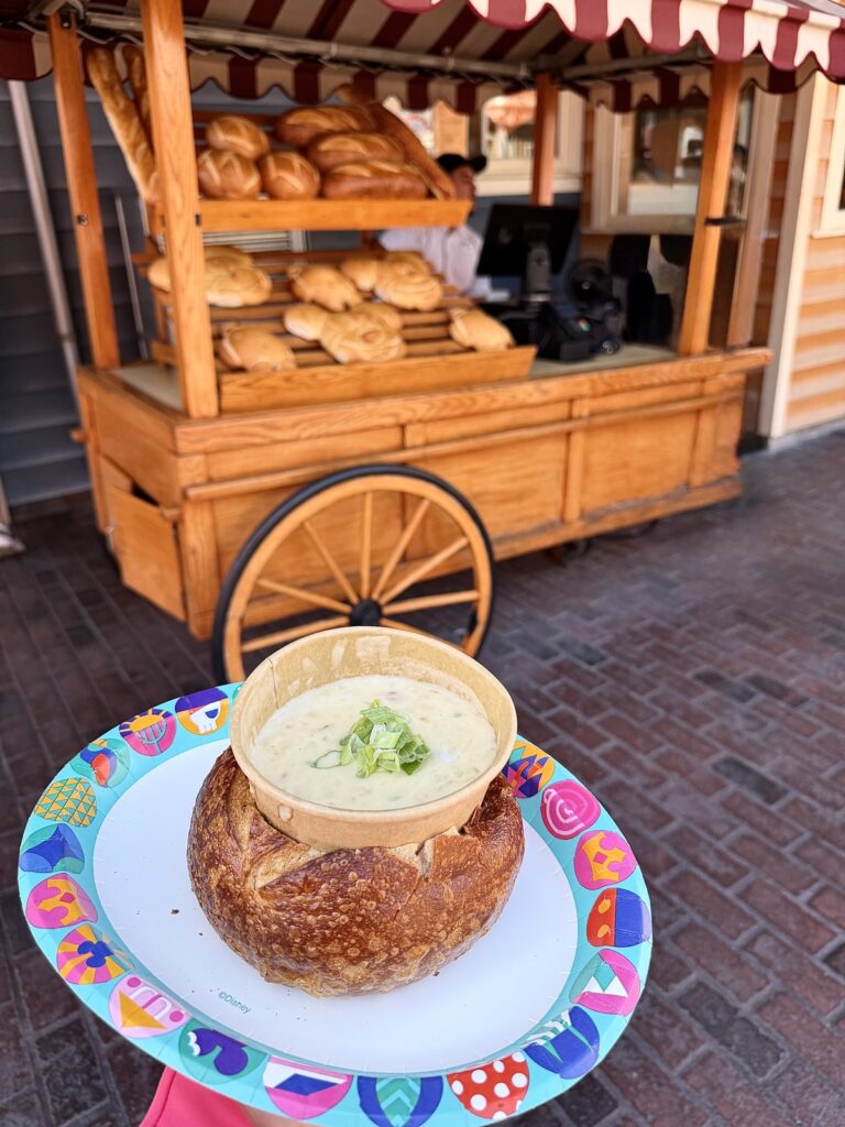 A picture of clam chowder and bread bowl from Aunt Cass in Disneyland, California Adventure, with the wagon where you can buy sourdough bread behind it.